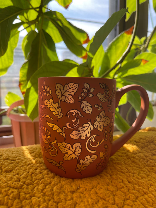 Terracotta mug with leaf pattern on a yellow surface with green plants in the background