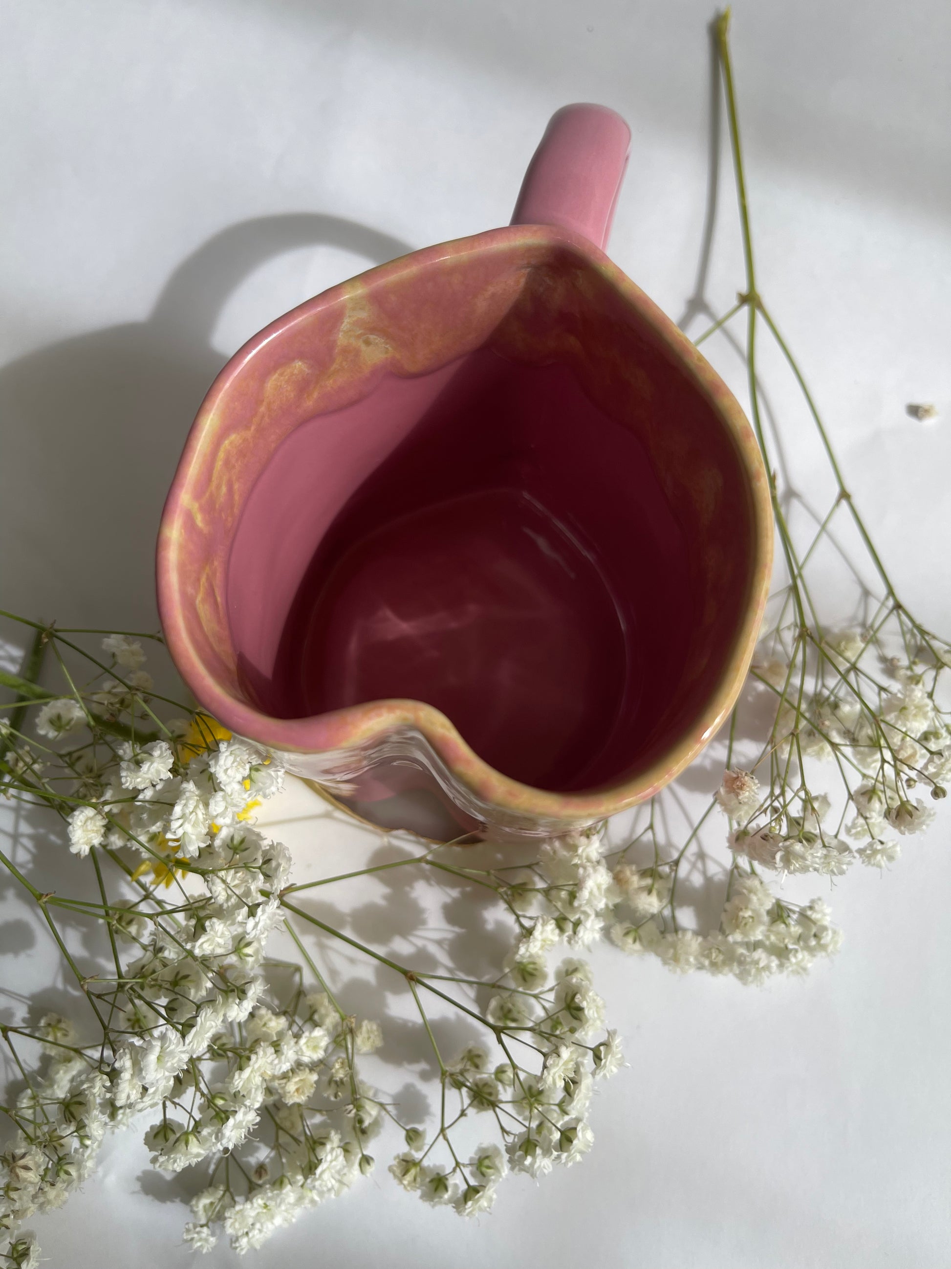Pink ceramic pitcher with a handle on a white surface with small white flowers around it.