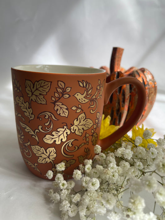 Brown mug with leaf pattern on a white surface with flowers and a pumpkin.
