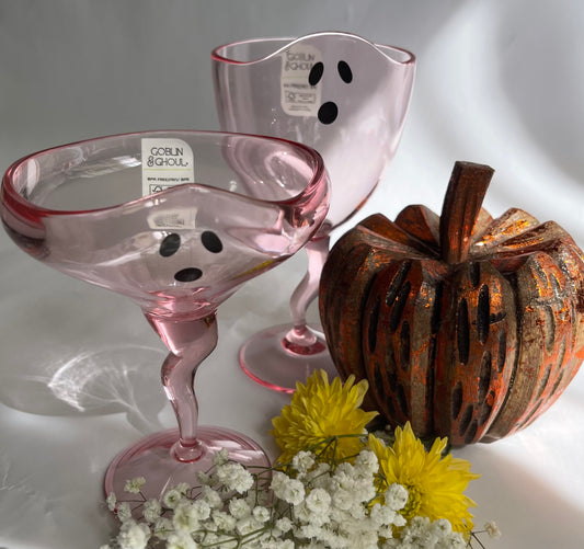 Pink ghost-shaped glasses with black polka dots, a decorative pumpkin, and flowers on a reflective surface.