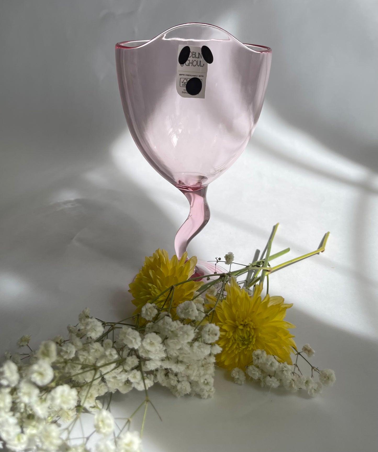 Pink glass goblet with a label on a white surface with yellow and white flowers.