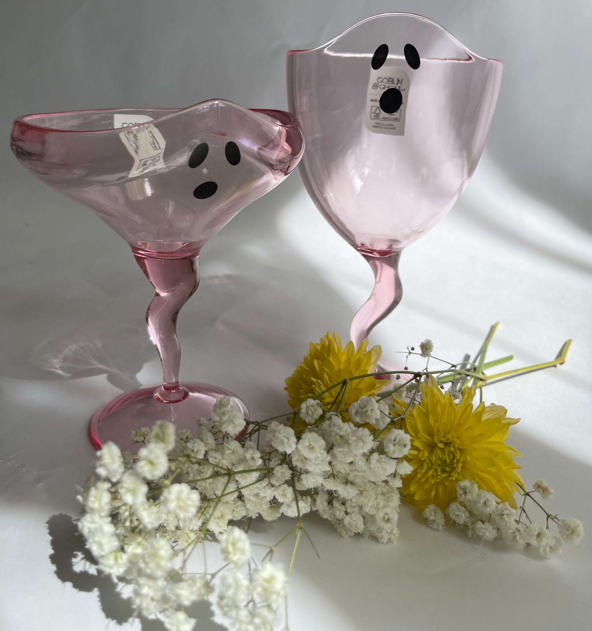 Two pink ghost-shaped glasses with black dots on a white surface with flowers.